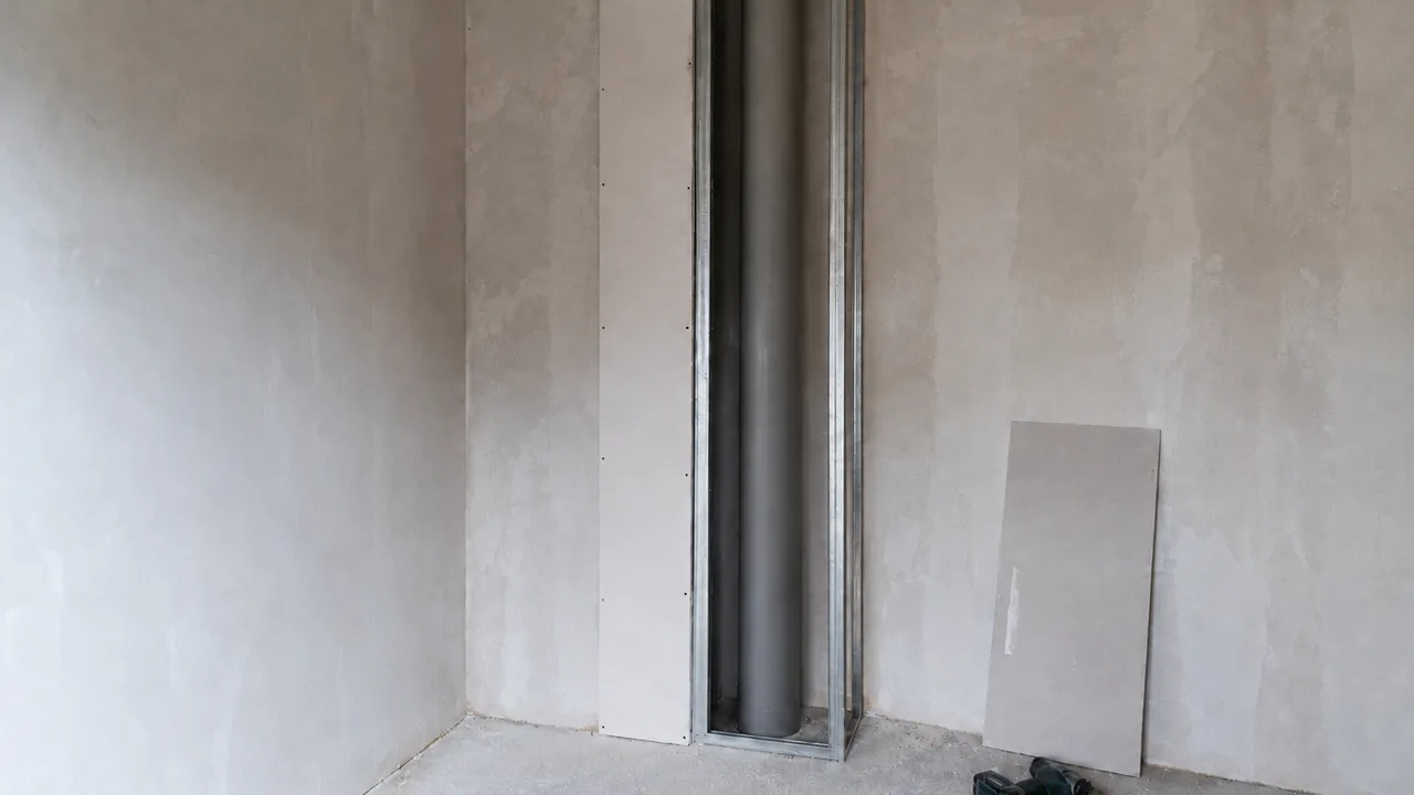 Canalisation verticale en PVC gris courant du sol au plafond dans l'angle d'une pièce en rénovation, entourée de l'ossature métallique de son coffrage dont la joue de gauche est déjà plaquée, le métal ne touchant le tuyau nulle part
