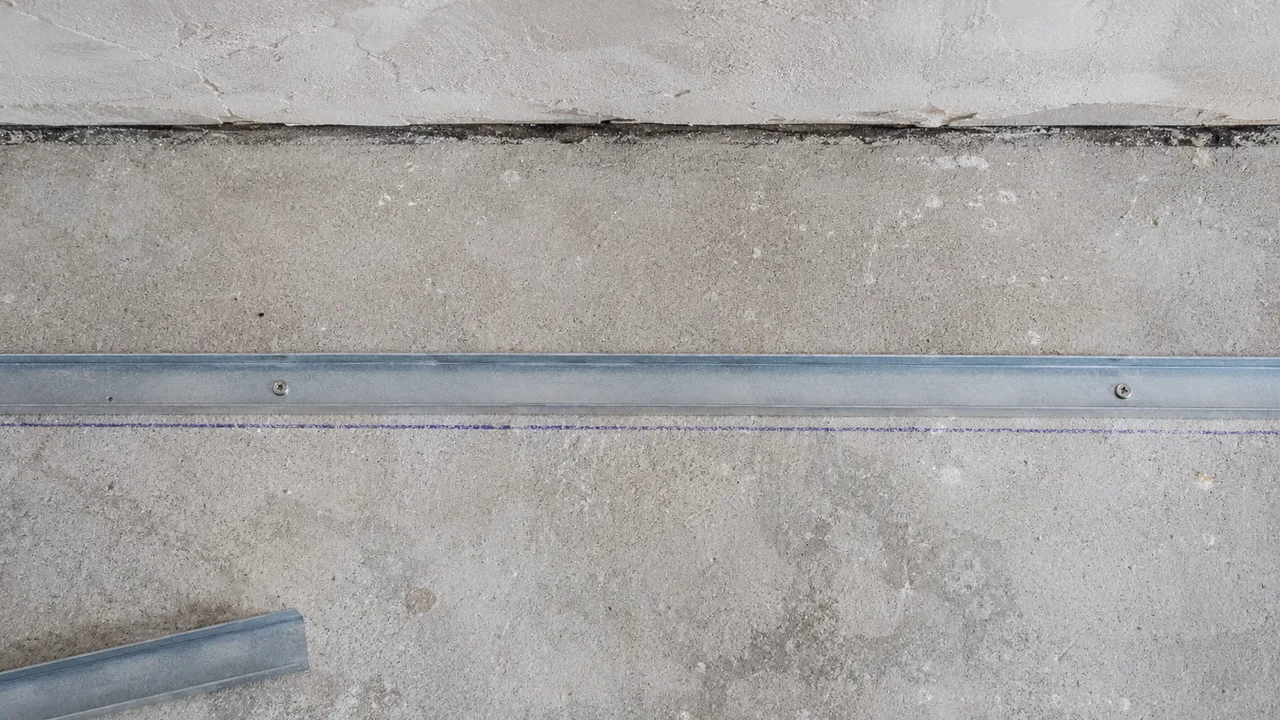 Rail métallique bas vissé à plat sur une dalle de béton, parallèle au pied du mur, avec la bande de dalle nue qui sépare le rail du mur et un trait de tracé bleu au cordeau