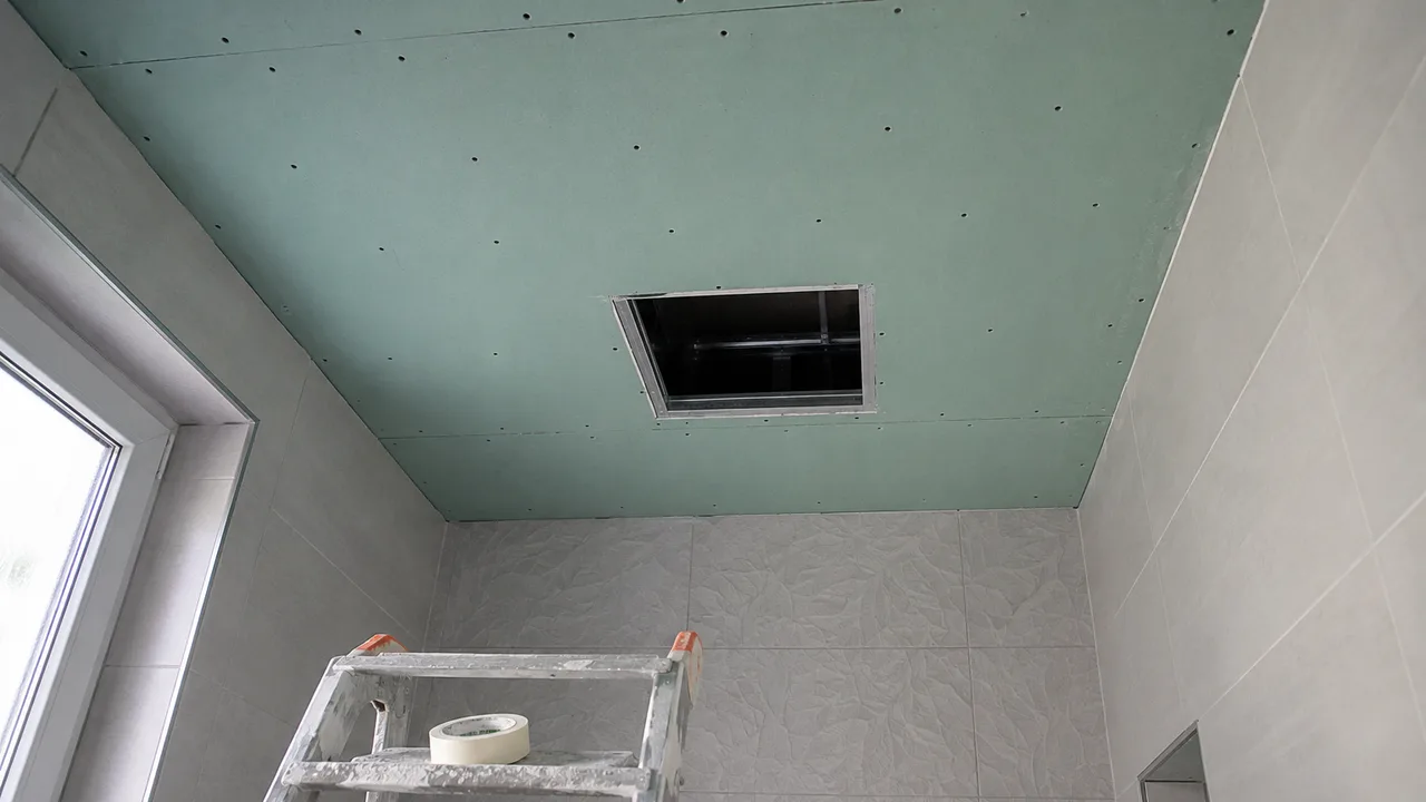 Plafond de salle de bain en cours de fermeture vu depuis le sol : plaques de plâtre hydrofuges vertes vissées sur leur ossature, têtes de vis et joints visibles, et ouverture rectangulaire découpée pour la future trappe de visite
