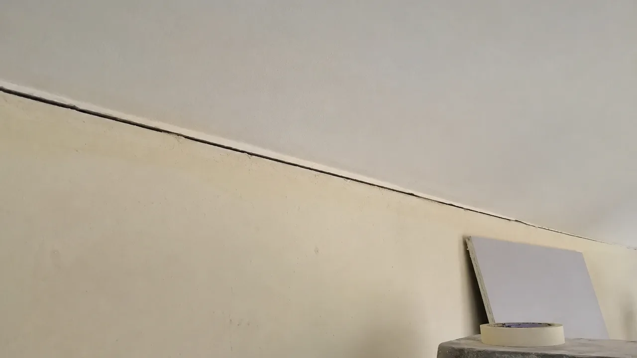 Jonction entre le haut d'un mur peint et le plafond d'un séjour en rénovation : la liaison s'est ouverte en un trait sombre continu qui suit l'arête sur toute sa longueur, bordé d'un liseré de peinture soulevée côté plafond