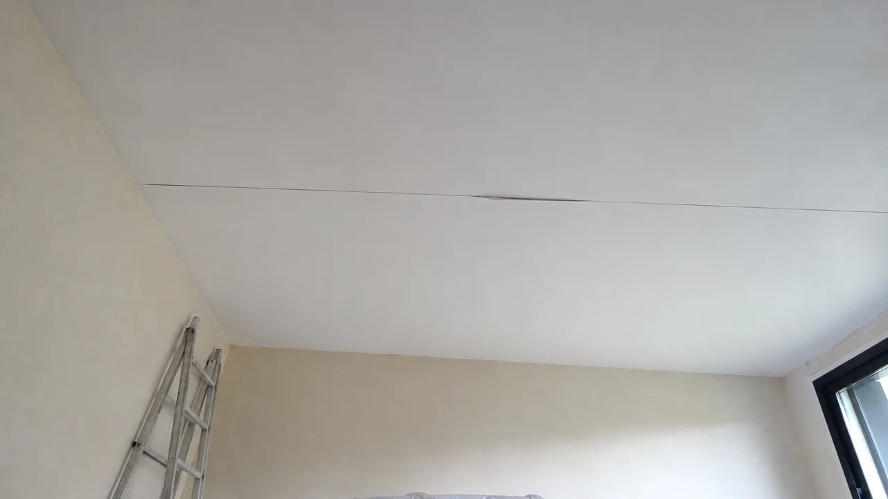 Fissure fine et rectiligne courant dans l'axe d'un joint au plafond d'une piece videe pour travaux, avec le papier de la bande a joint souleve sur une portion de sa longueur et un escabeau replie contre le mur