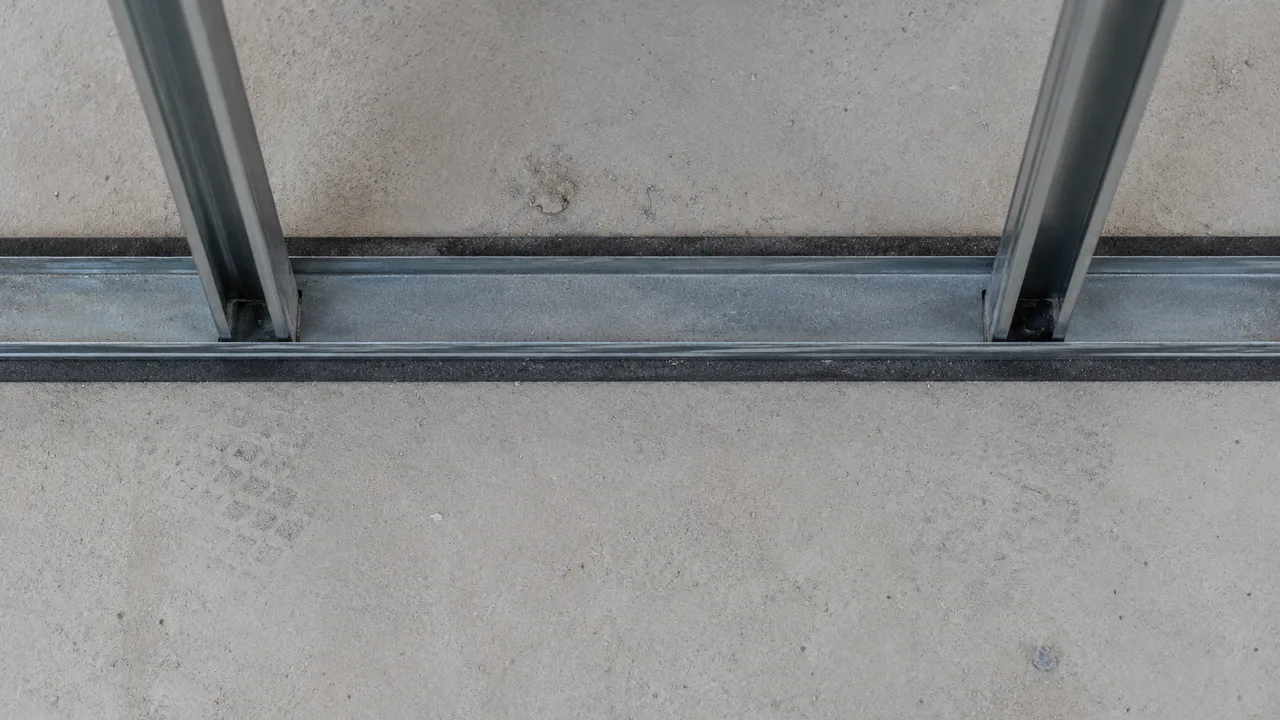 Rail métallique vu du dessus, posé sur une bande résiliente sombre qui dépasse de chaque côté sur toute sa longueur, avec deux montants engagés dedans sur une dalle de béton