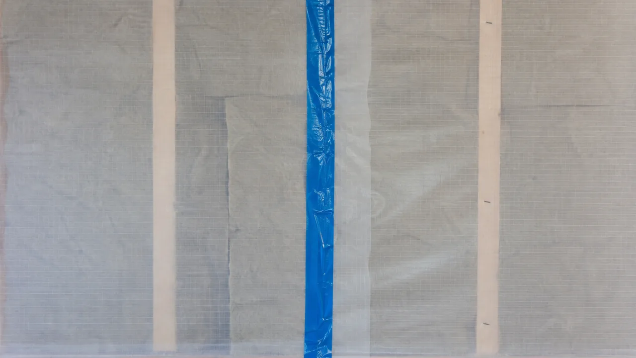 Recouvrement de deux lés de membrane frein-vapeur sur un mur d'ossature bois, fermé par une bande adhésive bleue marouflée à la main, laine minérale visible par transparence