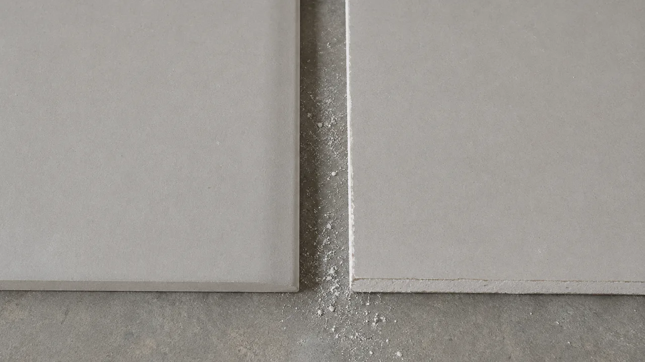 Deux morceaux de plaque de plâtre posés à plat bord à bord : à gauche un bord d'usine dont le carton se replie sur la tranche, à droite un bord de coupe dont l'âme de plâtre est à vif, avec ses éclats blancs et la poussière retombée entre les deux