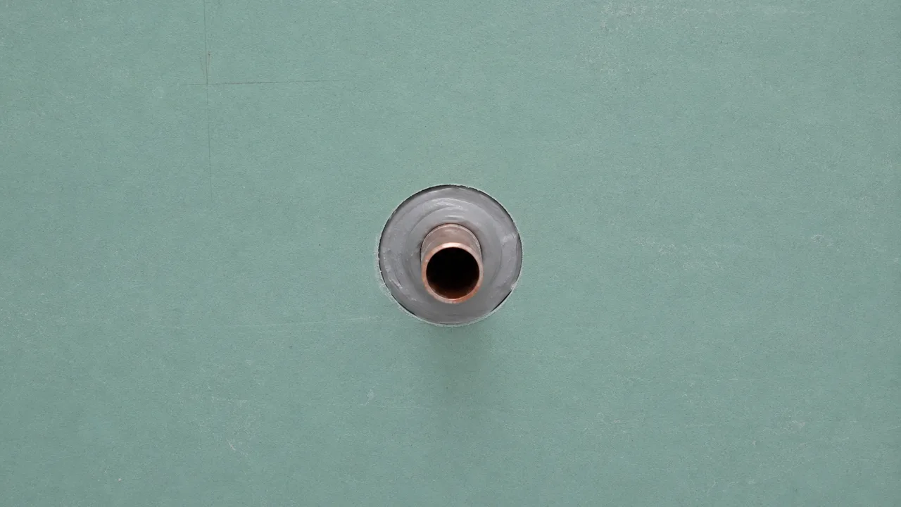 Sortie d'alimentation en cuivre traversant une plaque de plâtre hydrofuge verte, avec un cordon de mastic formant un anneau continu entre le tube et le bord de la découpe circulaire