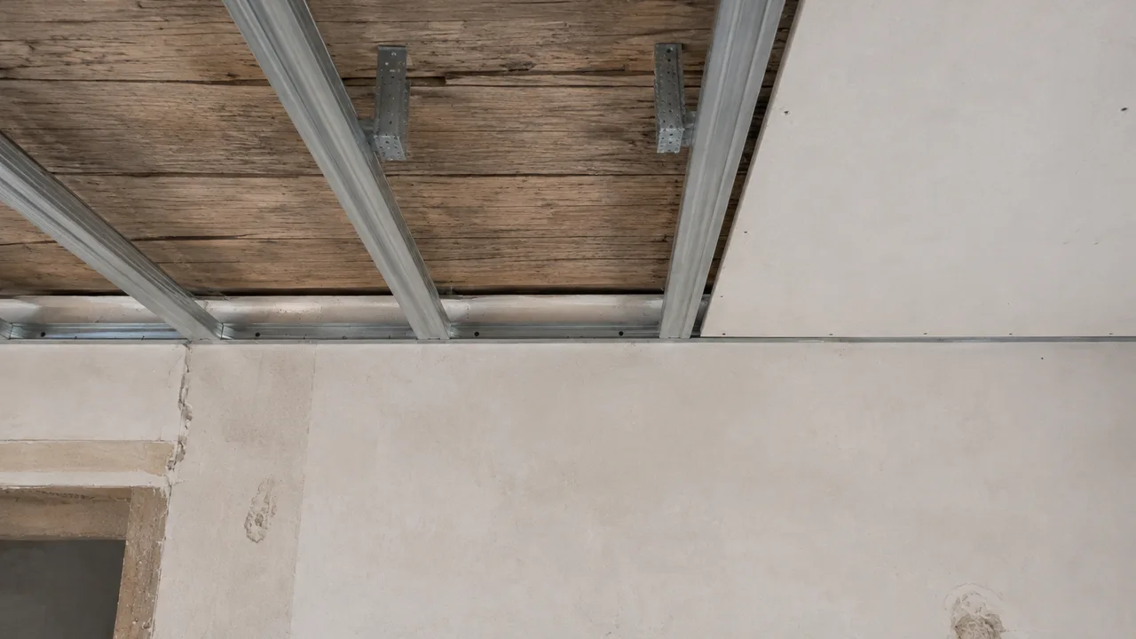 Extrémités des fourrures métalliques d'un plafond suspendu arrivant sur la cornière de rive vissée le long du mur, chaque profilé s'arrêtant avant la maçonnerie au lieu de venir buter contre elle
