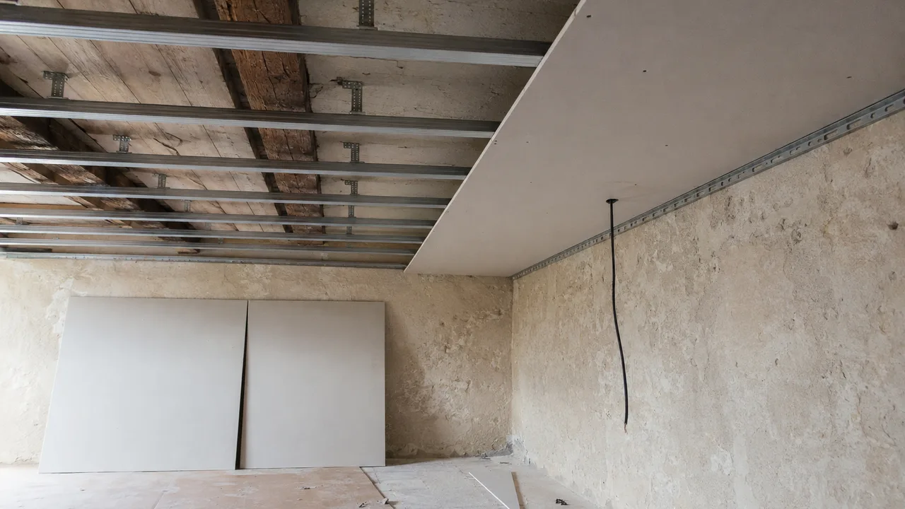 Plafond suspendu en cours de fermeture sous un vieux plancher de bois : les fourrures métalliques sont tenues à distance des poutres anciennes par des équerres vissées dans le bois, et la moitié droite du plafond est déjà couverte de plaques de plâtre