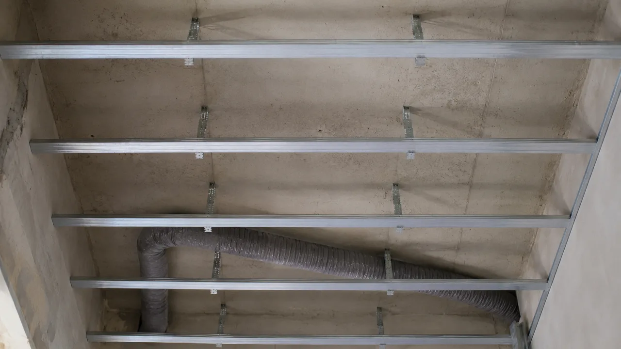 Ossature métallique de faux plafond vue par en dessous : les fourrures sont maintenues à distance de la dalle de béton brute par des équerres, et une gaine souple de ventilation serpente dans le vide ainsi ménagé