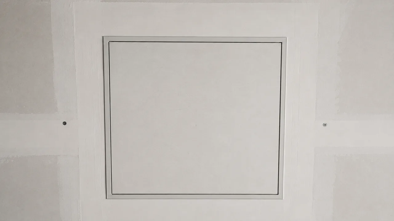Trappe de visite carrée fermée et affleurante dans un plafond en plaques de plâtre bandé, son fin cadre métallique dessinant un carré net sur la surface mate, entre deux têtes de vis alignées
